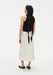 Rear three-quarter view of a woman against a plain white background, wearing a black sleeveless top with a long tie detail draping down the back, paired with a cream-colored midi skirt and black pointed heels. Her dark shoulder-length hair is worn down, and small tattoos are visible on her arms and upper back.