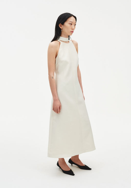 Side view of a model wearing a sleeveless cream midi dress with a high choker-style neckline and clean, minimal silhouette, paired with black pointed heels.