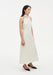 Side view of a model wearing a sleeveless cream midi dress with a high choker-style neckline and clean, minimal silhouette, paired with black pointed heels.