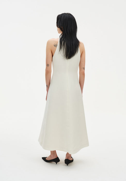 Back view of a model wearing a sleeveless cream midi dress with a clean A-line silhouette and racer-style back, paired with black low heels.