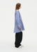 A woman stands in side profile against a plain white background, wearing a light blue striped long-sleeve shirt with a loose, flared hem, paired with black wide-leg trousers and light-colored shoes. Her straight light brown hair falls past her shoulders, and she maintains a neutral expression.
