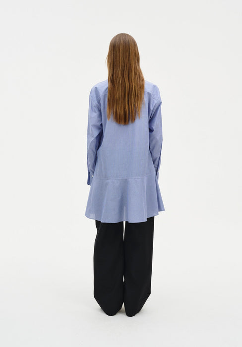 Rear view of a woman standing against a plain white background, wearing a light blue long-sleeve tunic-style top with subtle vertical texture and a flared hem, paired with loose black wide-leg trousers. Her straight light brown hair falls down her back.