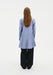 Rear view of a woman standing against a plain white background, wearing a light blue long-sleeve tunic-style top with subtle vertical texture and a flared hem, paired with loose black wide-leg trousers. Her straight light brown hair falls down her back.