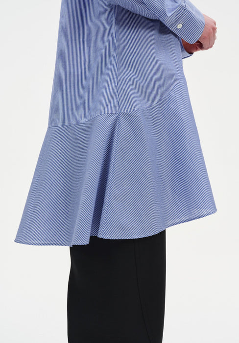 Close-up of a light blue long-sleeve tunic with fine vertical stripes, highlighting a curved seam and flared hem detail, worn over black wide-leg trousers.