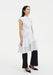 Side view of a model wearing a sleeveless white button-down tunic with a pointed collar and layered ruffled hem, styled over black cropped trousers and paired with black pointed heels.
