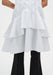 Close-up of a white sleeveless button-down tunic highlighting the layered, flared hem with soft ruffle tiers, worn over black trousers.