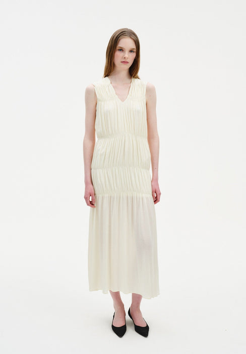 A woman stands facing forward against a plain white background, wearing a sleeveless cream-colored midi dress with a V-neckline and gathered tiered detailing, paired with black pointed heels. Her straight light brown hair is worn down, and she has a neutral expression with arms relaxed at her sides.