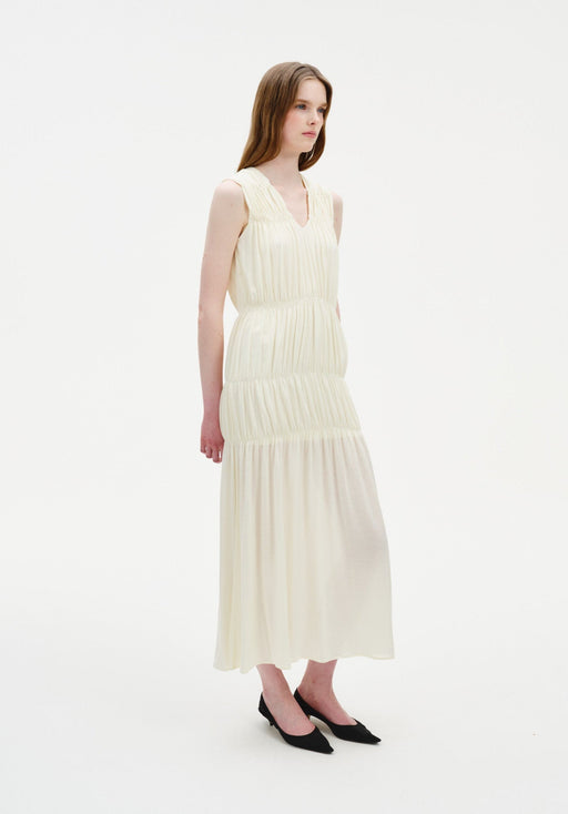 A woman stands in a three-quarter side pose against a plain white background, wearing a sleeveless cream-colored midi dress with gathered tiers and a softly flowing silhouette, paired with black pointed heels. Her straight light brown hair is worn down, and she looks slightly to the side with a neutral expression.