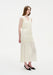 A woman stands in a three-quarter side pose against a plain white background, wearing a sleeveless cream-colored midi dress with gathered tiers and a softly flowing silhouette, paired with black pointed heels. Her straight light brown hair is worn down, and she looks slightly to the side with a neutral expression.