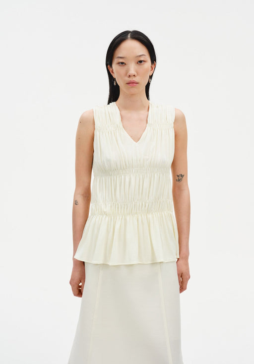 Front view of a sleeveless cream blouse with a V-neckline, gathered shirring, and peplum hem, paired with a matching skirt.